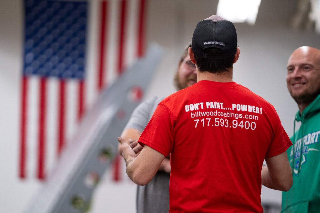 Biltwood Powder Coating team members talking in the shop, featuring branded shirt with company website and phone number.