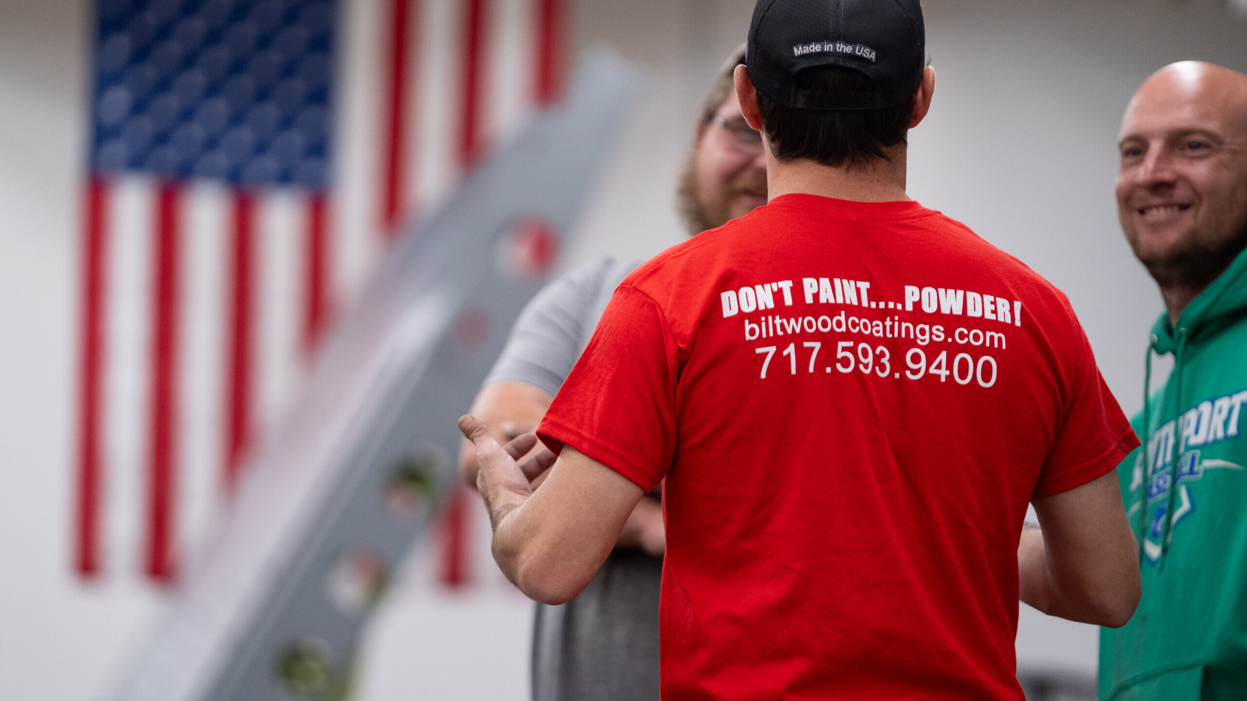 Biltwood Powder Coating team members talking in the shop, featuring branded shirt with company website and phone number.